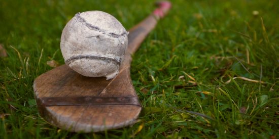Picture of a white sliotar sitting on top of a weathered hurl on grass