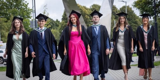 Six graduates walking in a row, girls in a pink dress in the middle