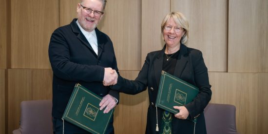 Tall man with glasses in a black jacket, holding a green folder, shakes hands with a woman in black, also holding a green folder