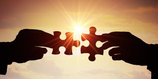 2 hands, each holding a jigsaw piece meeting in the middle. Background includes a setting sun. 