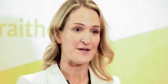 A blonde haired woman in a white blazer speaking to camera - yellow and white backdrop