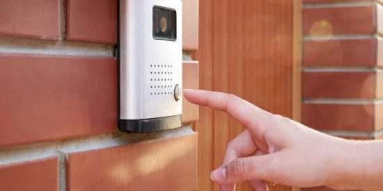 Person pushing button on white smart doorbell, red brick house, brown door