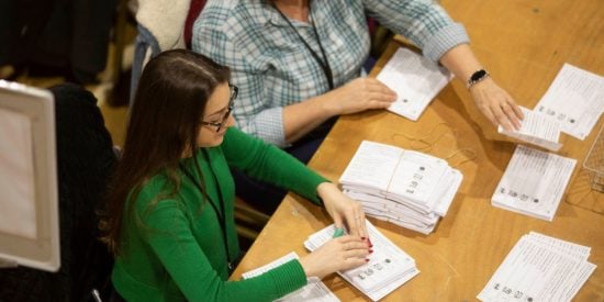 2 women - women on the left with a green top, sitting at a desk counting ballot papers, woman on the right wearing a blue/white blouse, also counting ballot papers. 