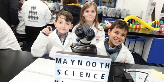 Two boys and a girl wearing white coats stand behind a microscope and sign reading Maynooth Science Night 2024