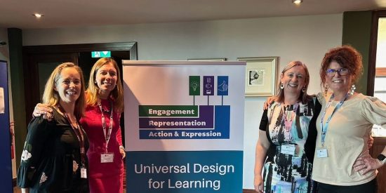 Four Women are standing on either side of the UDL International Symposium banner.