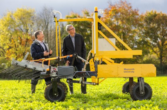 2 men standing in a field, behind a yellow seed robotic planter