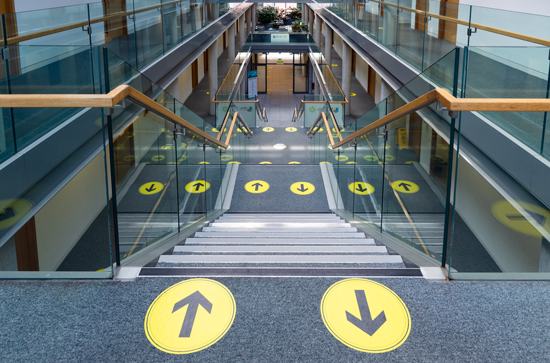 Social distancing signage on the stairs in Iontas showing which side to walk up and down. The stickers are black arrows on yellow circles