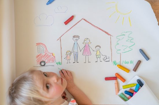 blonde hair child using crayons to draw house, with family and car