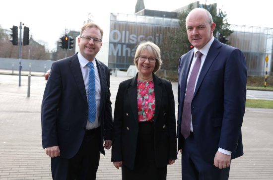 3 people posing for camera, in front of large building with font Ollscoil Mha Nuad. Man to left in dark suit, white shirt, blue tie, glasses and brown hair. Women in middle dark suit with pink, blue and green floral top, with glasses and blonde hair. Man to the right, navy blue suit with lilac shirt and purple tie. Tall man