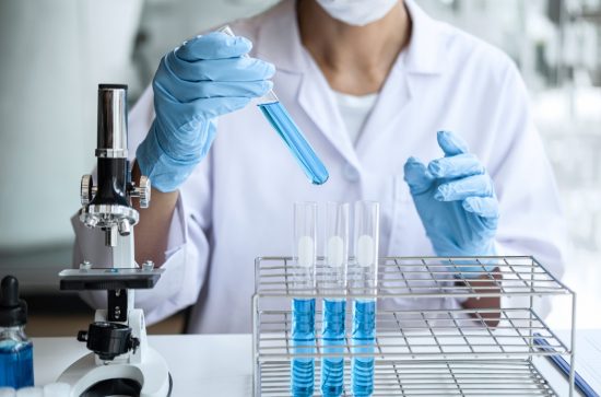 Scientist in a white lab coat and blue gloves, holding a test tube with blue liquid beside a microscope