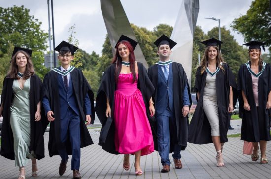 Six graduates walking in a row, girls in a pink dress in the middle