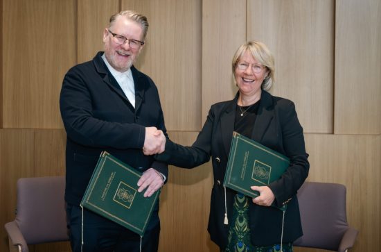 Tall man with glasses in a black jacket, holding a green folder, shakes hands with a woman in black, also holding a green folder