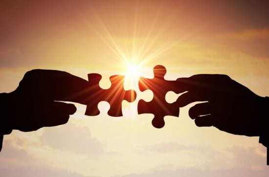 2 hands, each holding a jigsaw piece meeting in the middle. Background includes a setting sun. 