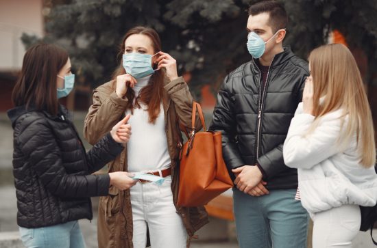 3 women and 1 man talking to each other wearing covid masks. Black jackets. blue jeans, white jacket and brown jacket 