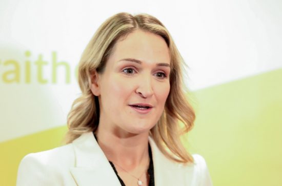 A blonde haired woman in a white blazer speaking to camera - yellow and white backdrop