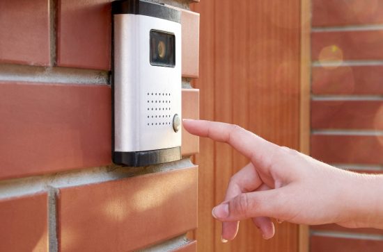 Person pushing button on white smart doorbell, red brick house, brown door