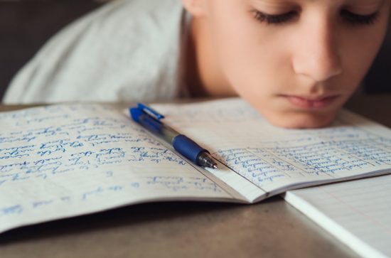 Child with sad face and head resting on table. Book with writing and a pen